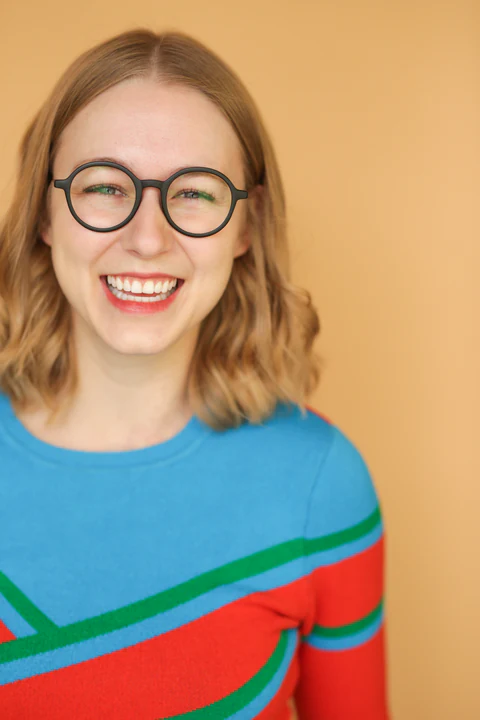 Katie Peabody headshot — round glasses, striped colour-block sweater, broad laugh