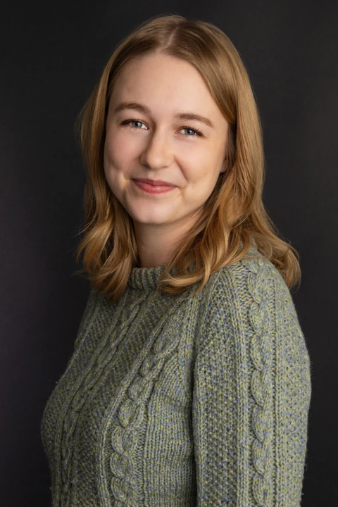 Katie Peabody headshot — green cable-knit sweater, dark background, three-quarter turn