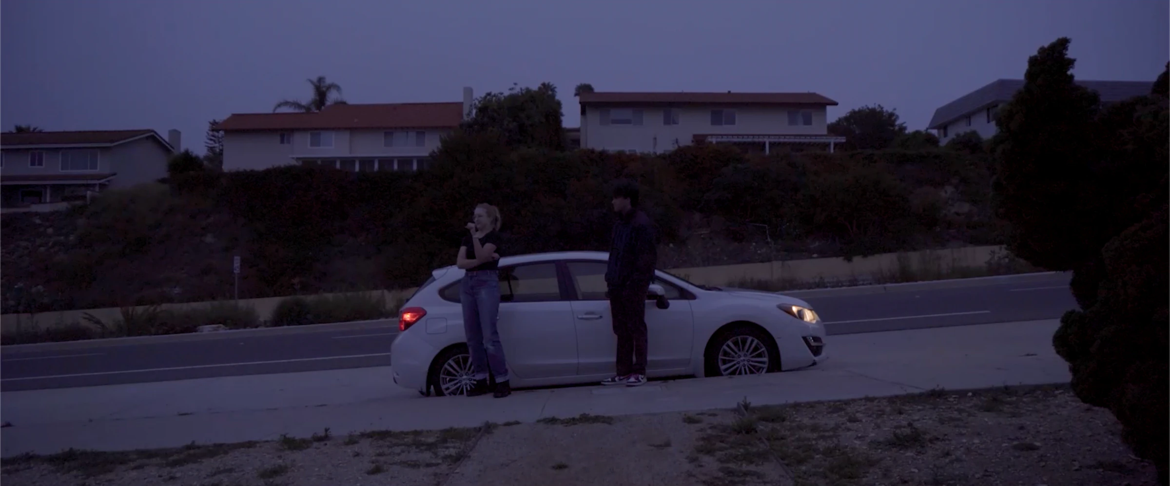 Katie Peabody as Aura in the film Sapphire Kingshit, two figures beside a car on a coastal street
